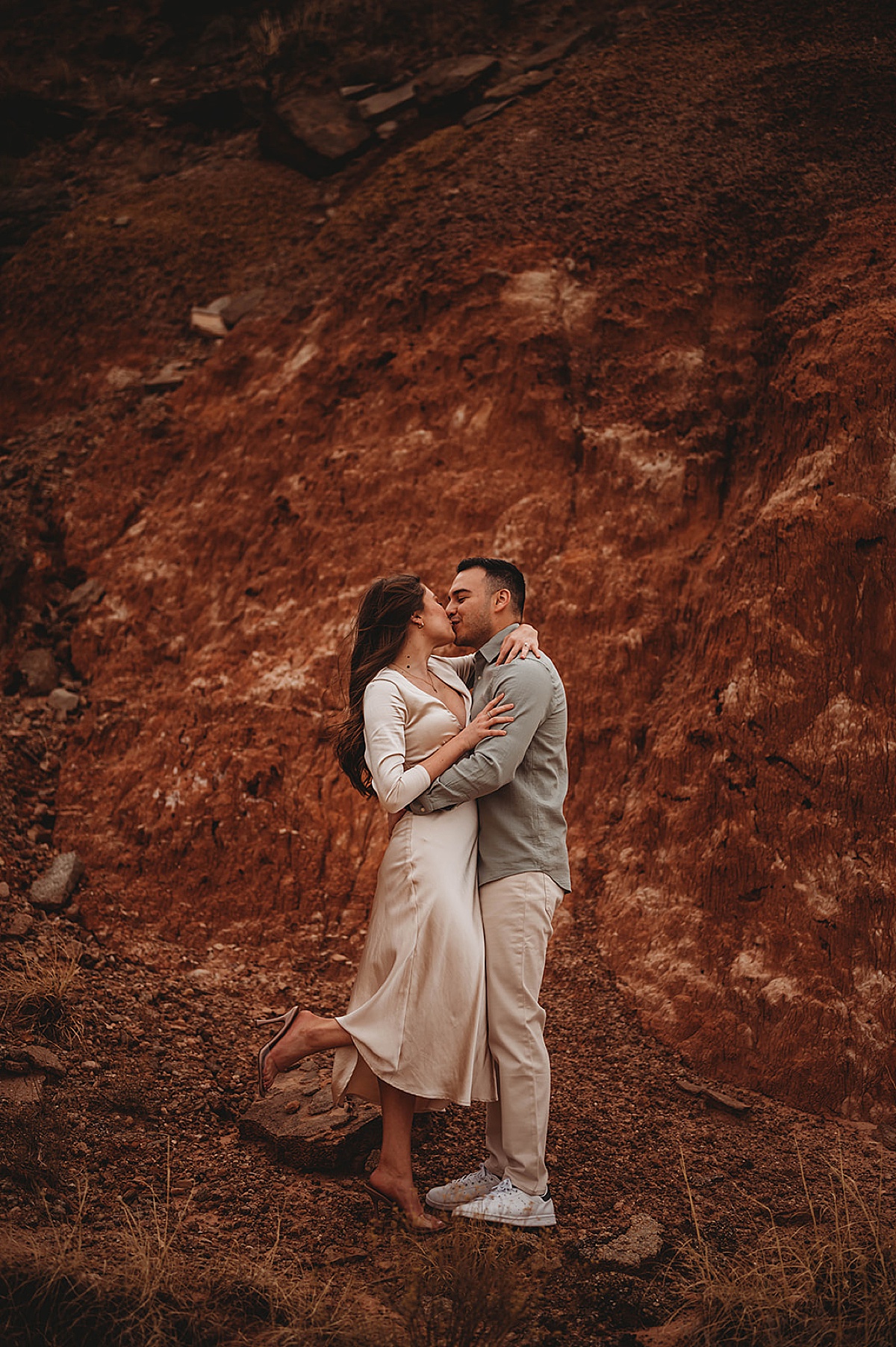 man and woman in silk dress kiss in front of red rock cliff at engagement shoot by Three Feather Photo Co