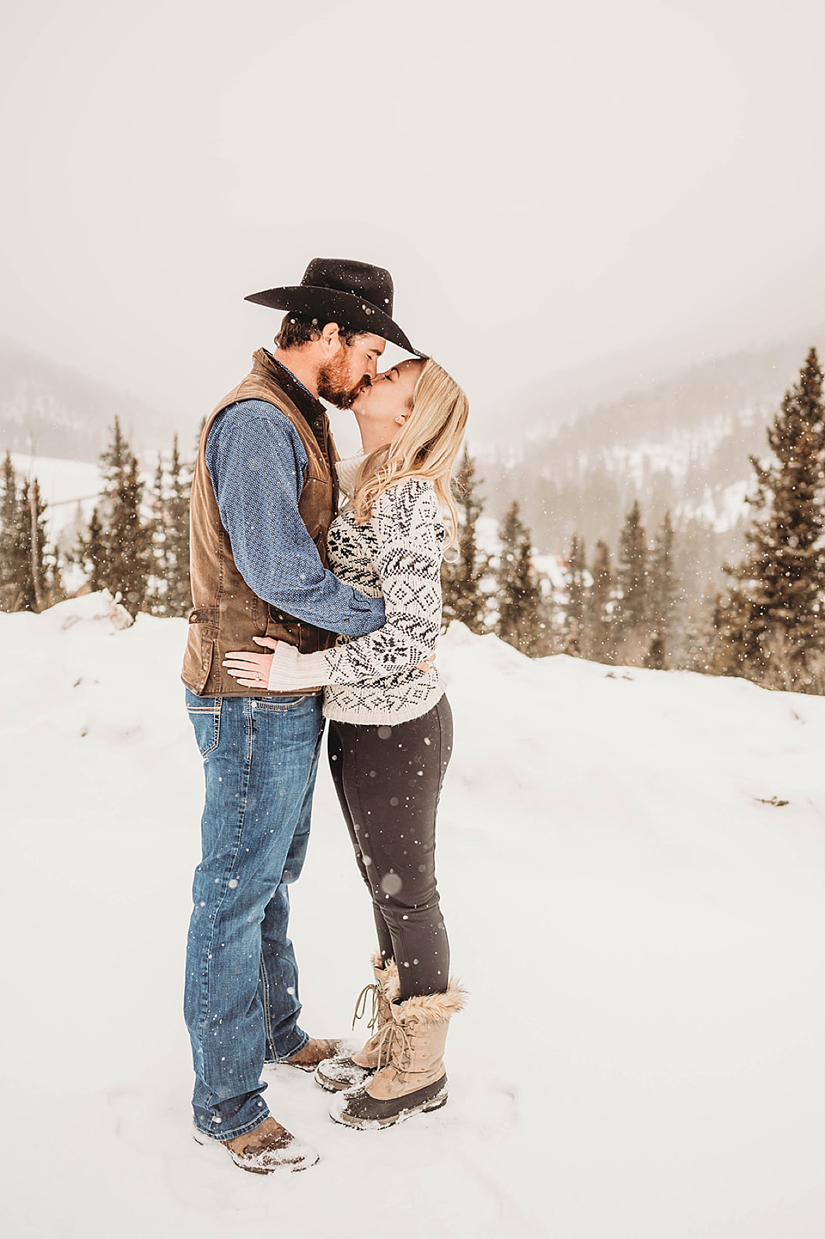 couple in sweater and cowboy hat kisses as the snow falls in western winter engagement shoot