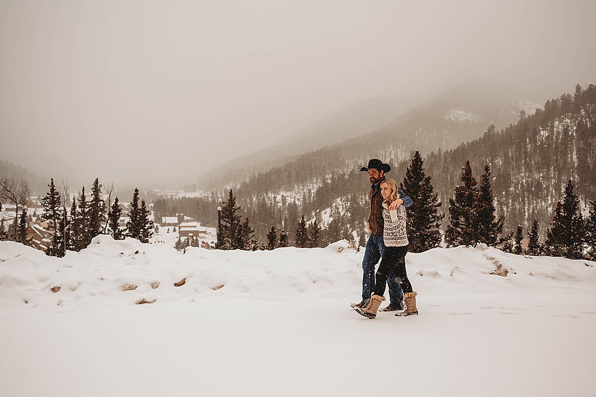 couple in snow boots and sweater walk through forest in red river NM western winter engagement shoot