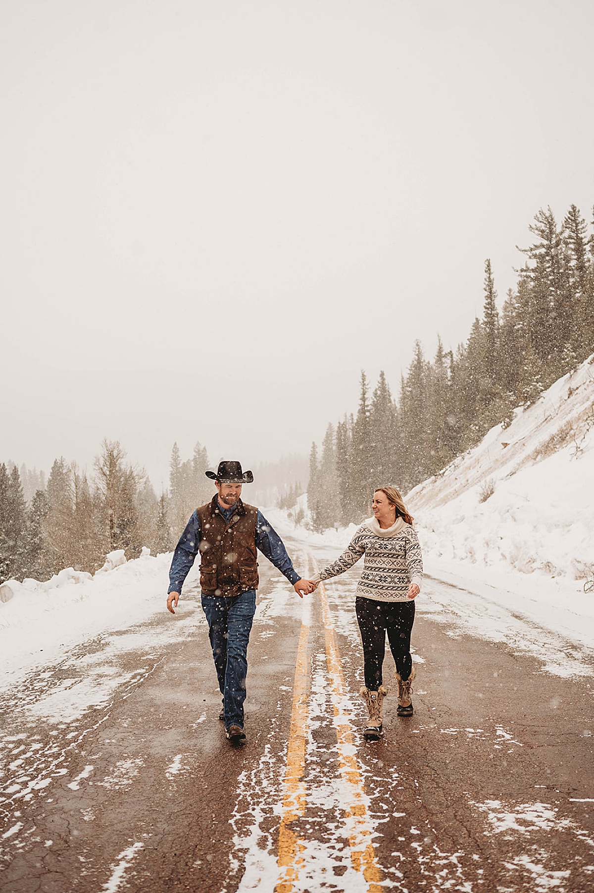 couple in sweater and vest walk down mountain road in the snow during engagement shoot by Three Feather Photo Co