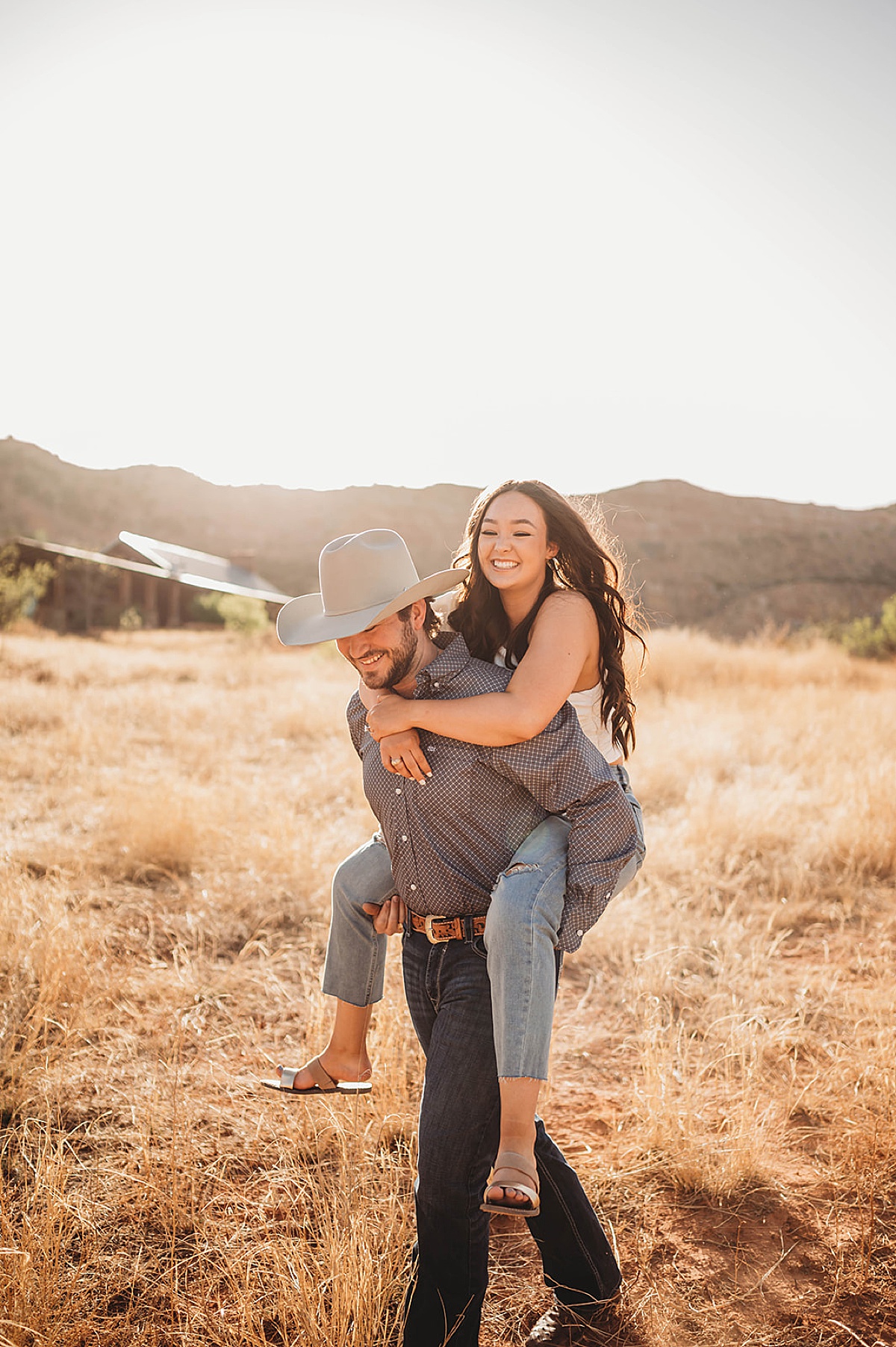 cute couple pose piggy back in field during golden summer engagement shoot