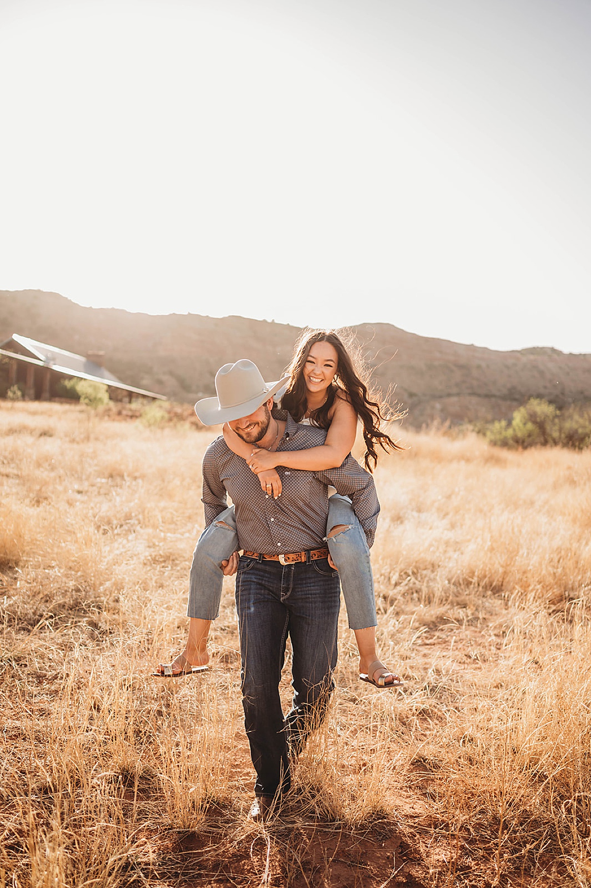 man in cowboy hat gives his fiancee a piggy back ride during golden summer engagement shoot