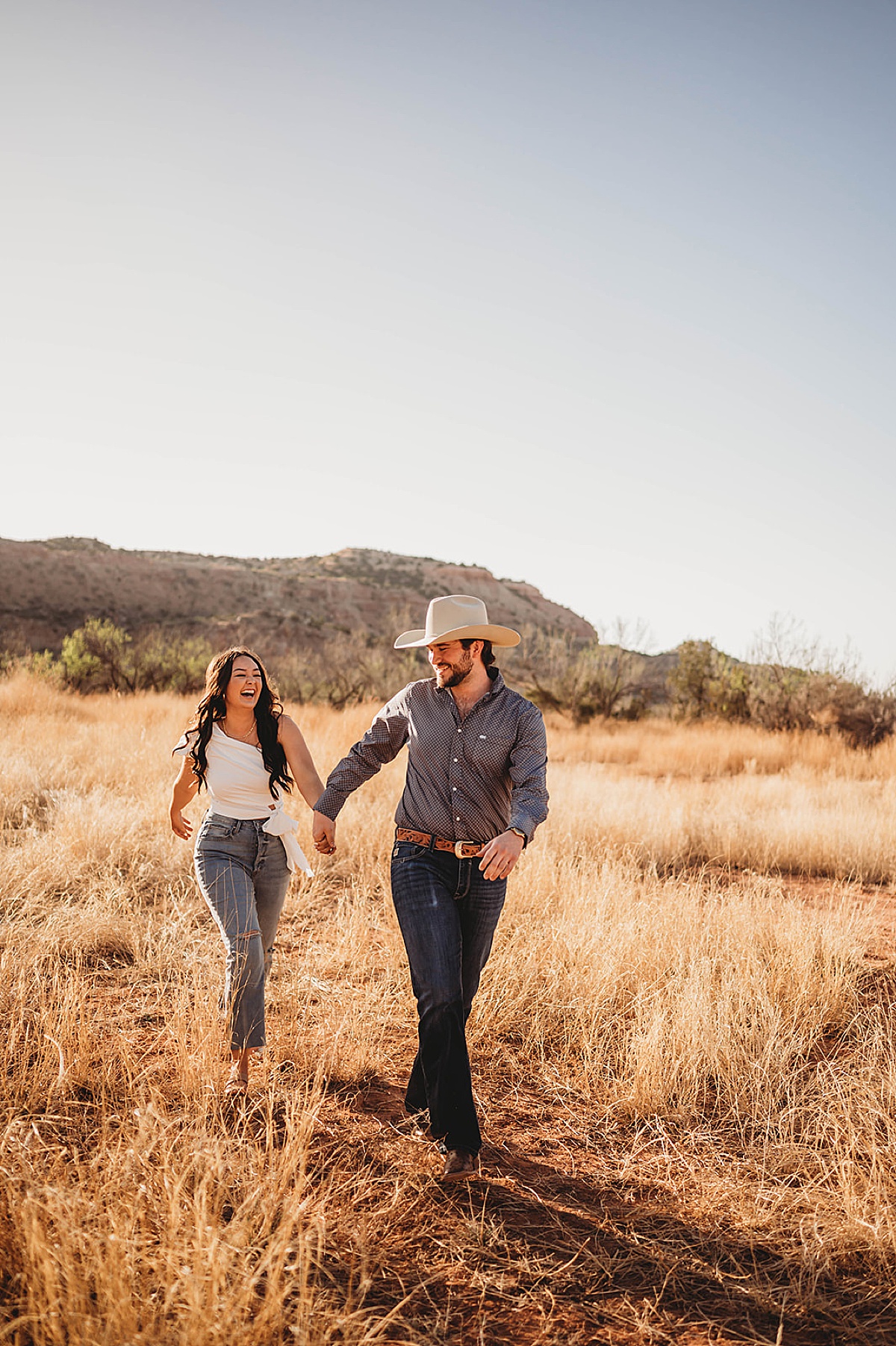 couple in nice casual clothes walk through field during golden summer engagement shoot