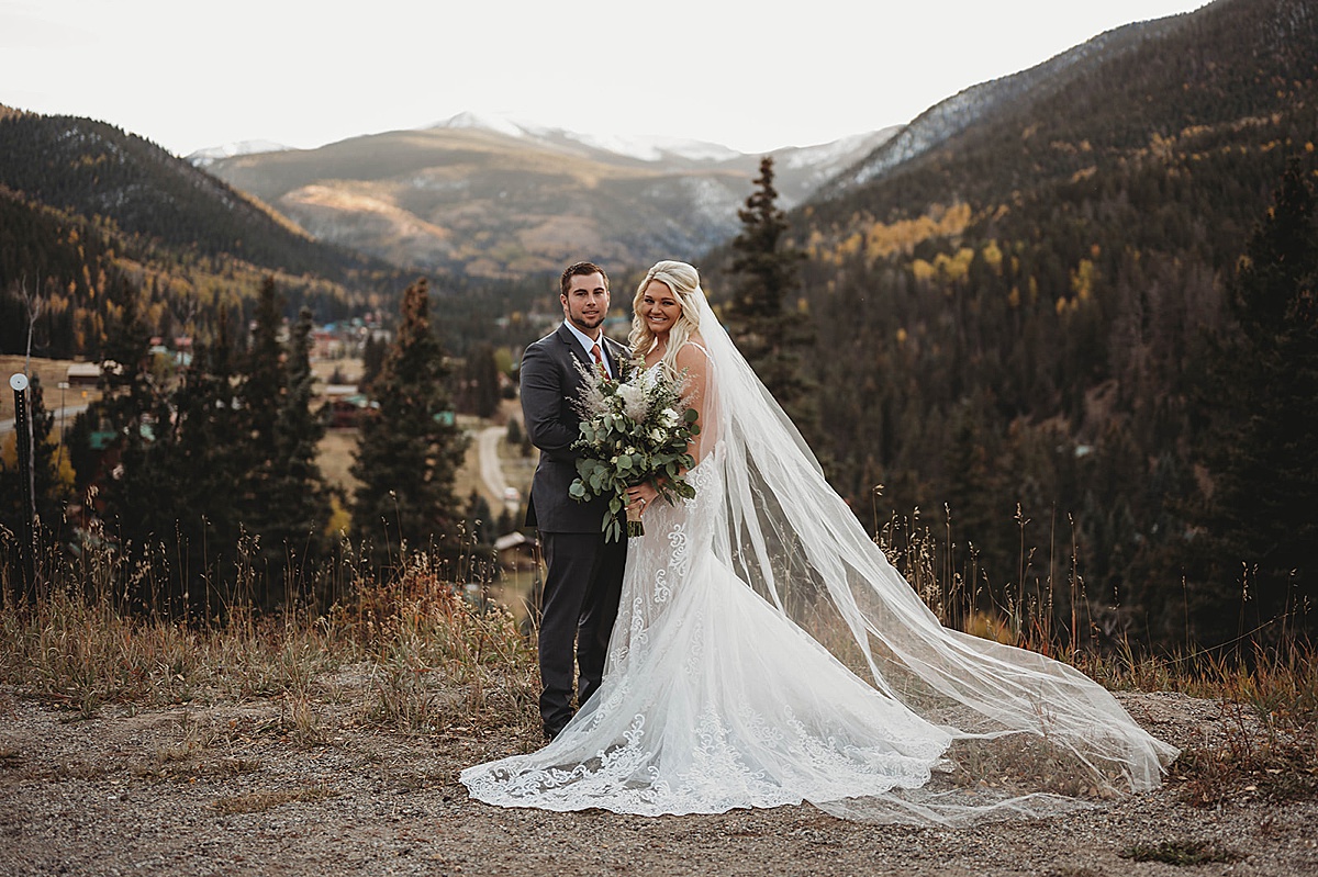 Bride in gown with train and veil poses with groom in front of autumn mountain view after wedding shot by Three Feather Photo Co