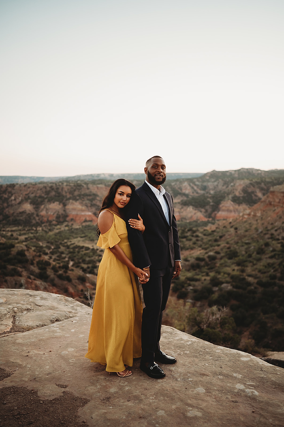 man in sharp suit and woman in yellow evening gown pose in front of dramatic view during engagement shoot by Three Feather Photo Co.