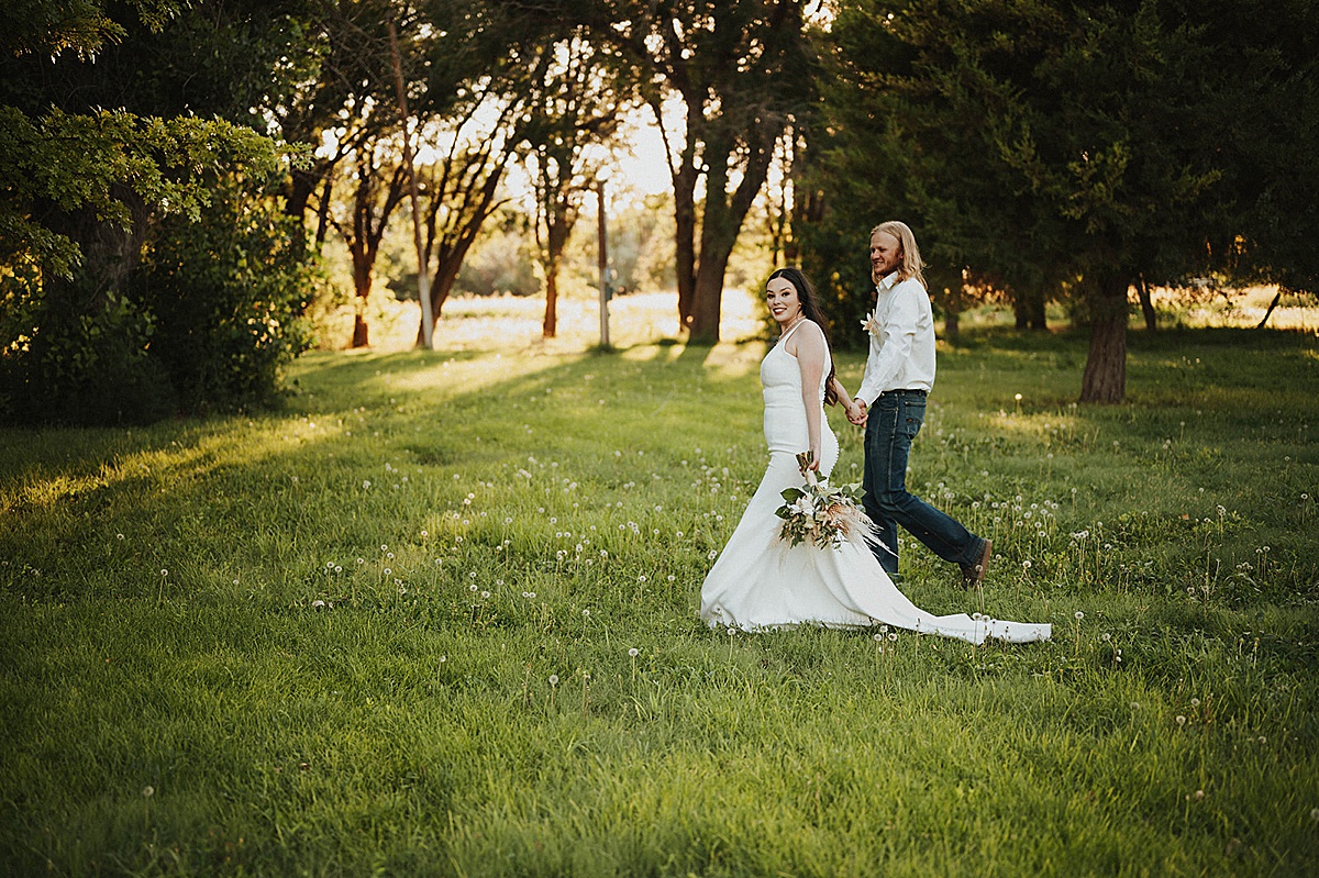 bride in minimalist chic gown and groom in classy western wear walk across field after sweet summer elopement