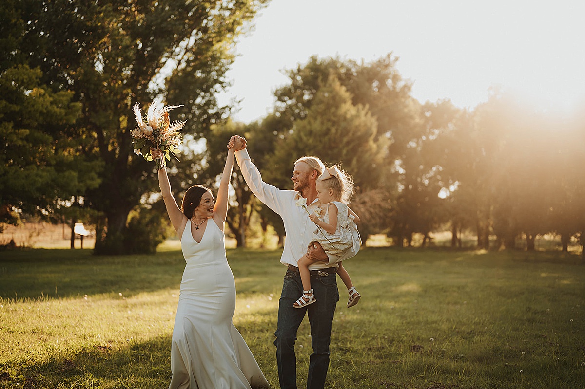 newlywed couple celebrate in golden hour field with their young daughter after wedding shot by Three Feather Photo Co
