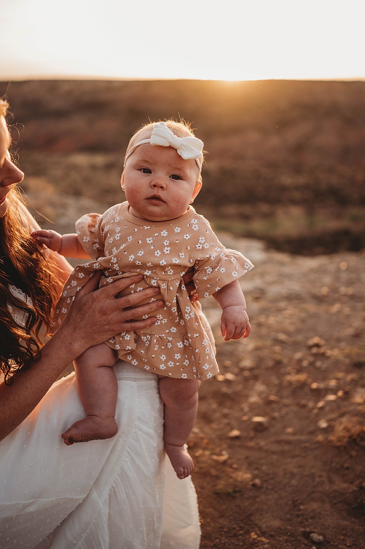 baby girl in flowered dress and headband with bow sits on mom's knee during shoot with three feather photo co