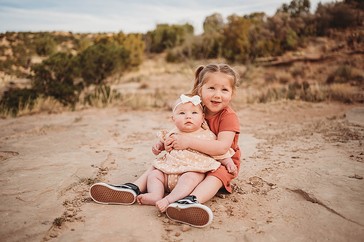 big sister in orange dress holds baby sister on her lap during cute outdoor canyon session with three feather photo co