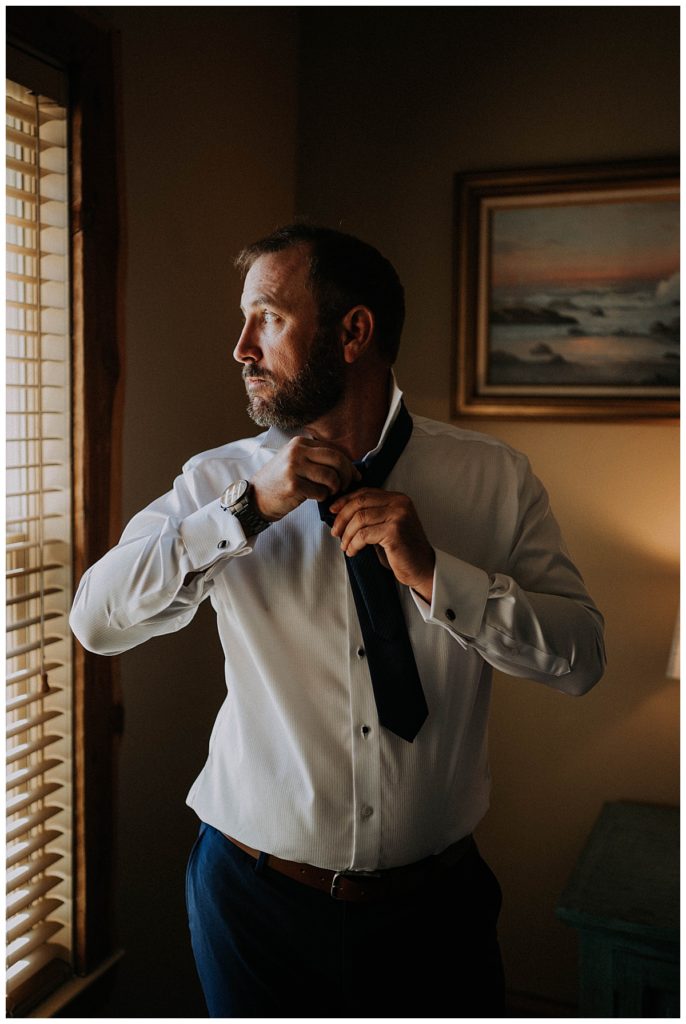 husband fixes tie while looking out window by Three Feather Photo