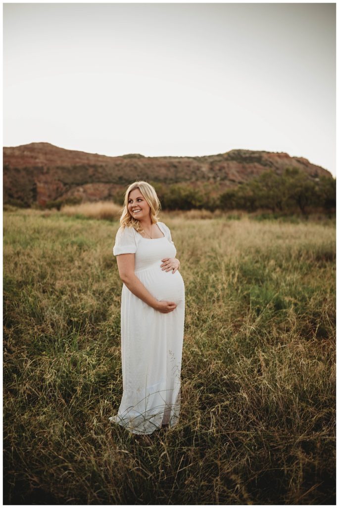 blonde woman in white dress holds belly by Three Feather Photo