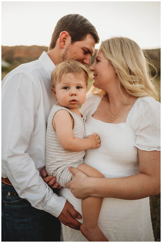 parents lean to kiss while holding child by Three Feather Photo