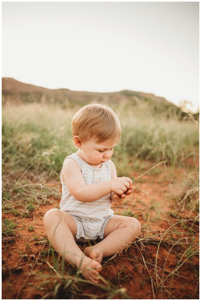 child plays with grass while sitting in dirt by Three Feather Photo